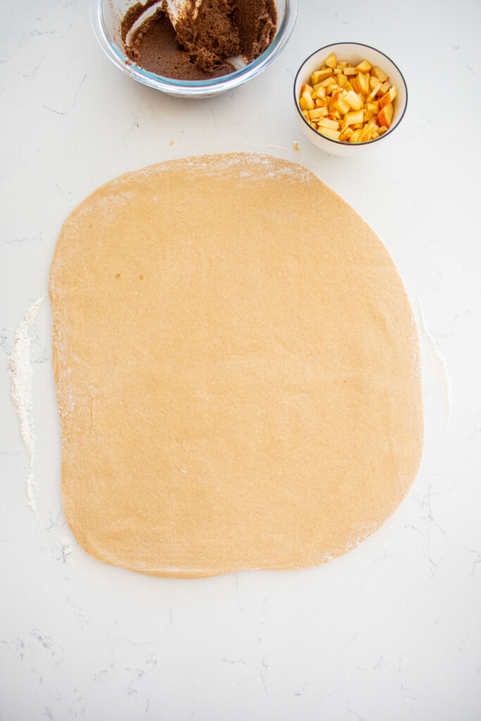 caramel apple pie cinnamon roll dough rolled out on a marble countertop with a bowl of chopped apples beside it.