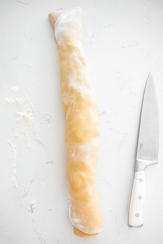 caramel apple pie cinnamon roll dough rolled into a log with a knife beside it on a marble counter.