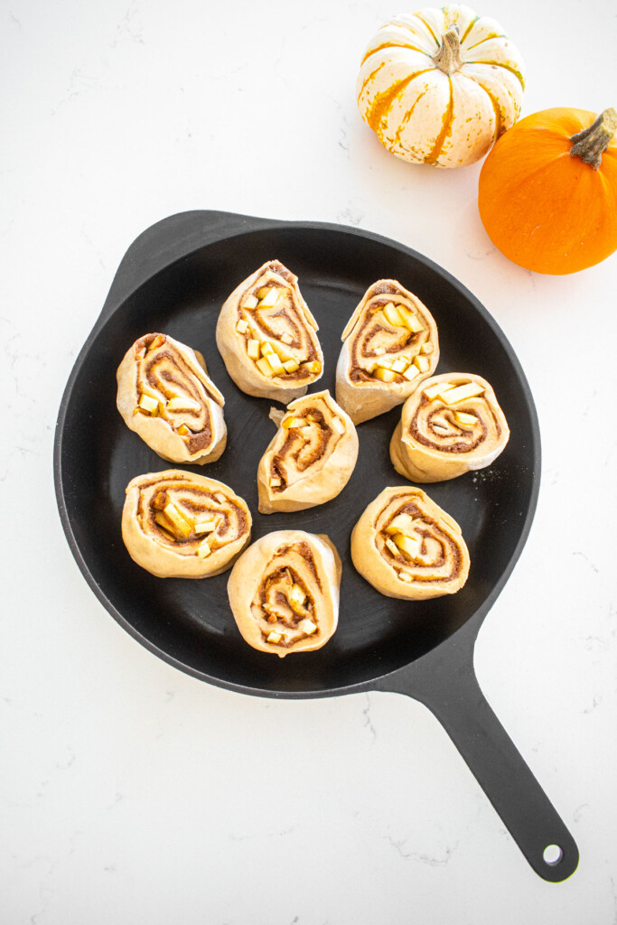 cast iron skillet with caramel apple pie cinnamon roll dough inside it on a marble counter with tiny pumpkins.