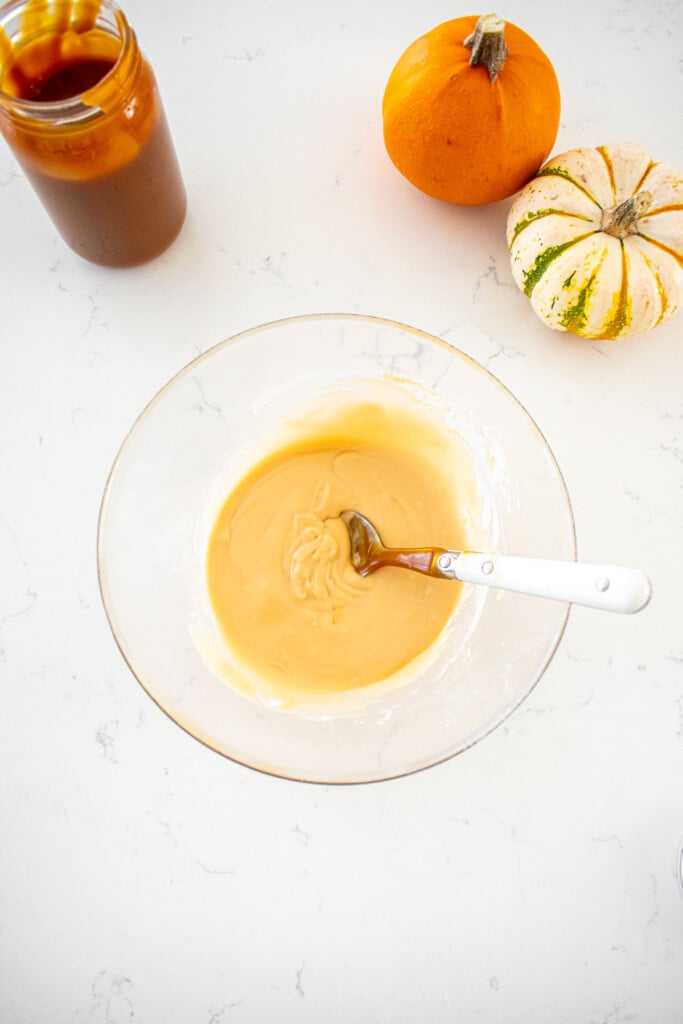 caramel glaze in a glass mixing bowl with a spoon on a marble counter with tiny pumpkins and a jar of caramel sauce.