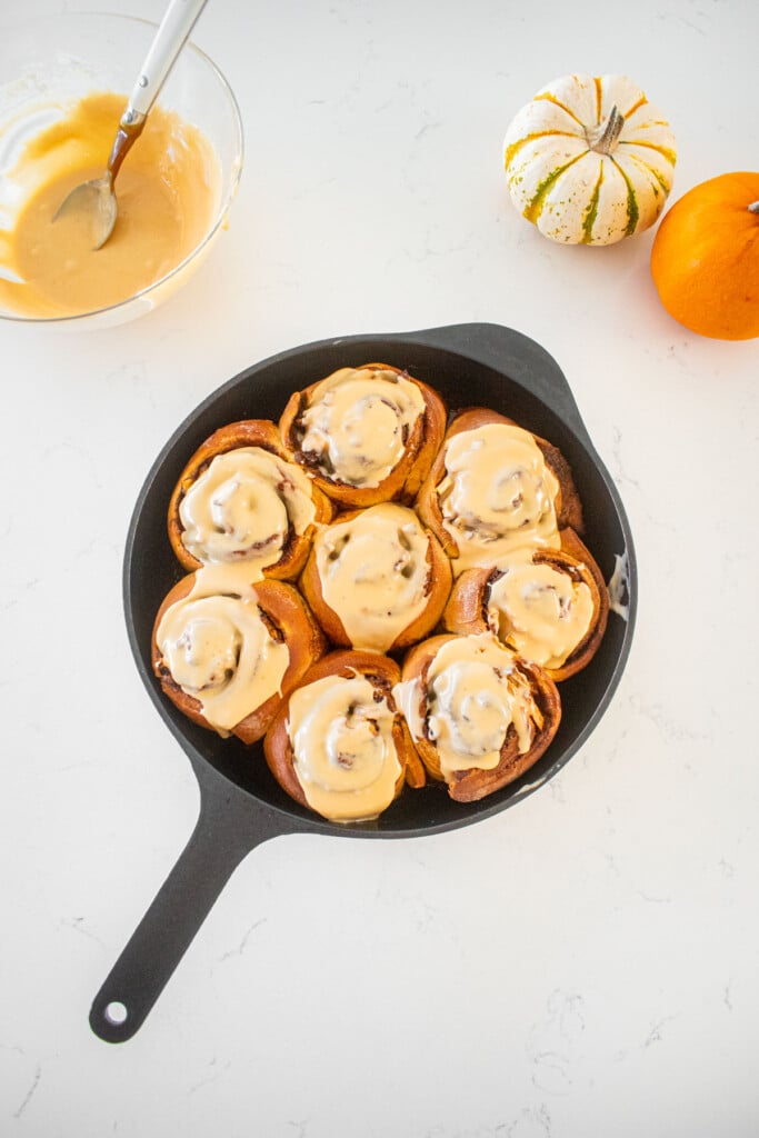 caramel apple pie cinnamon rolls with caramel glaze on top in a cast iron skillet on a marble counter with tiny pumpkins.