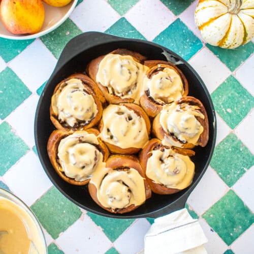 caramel apple pie cinnamon rolls in a cast iron skillet on a tile table with apples.