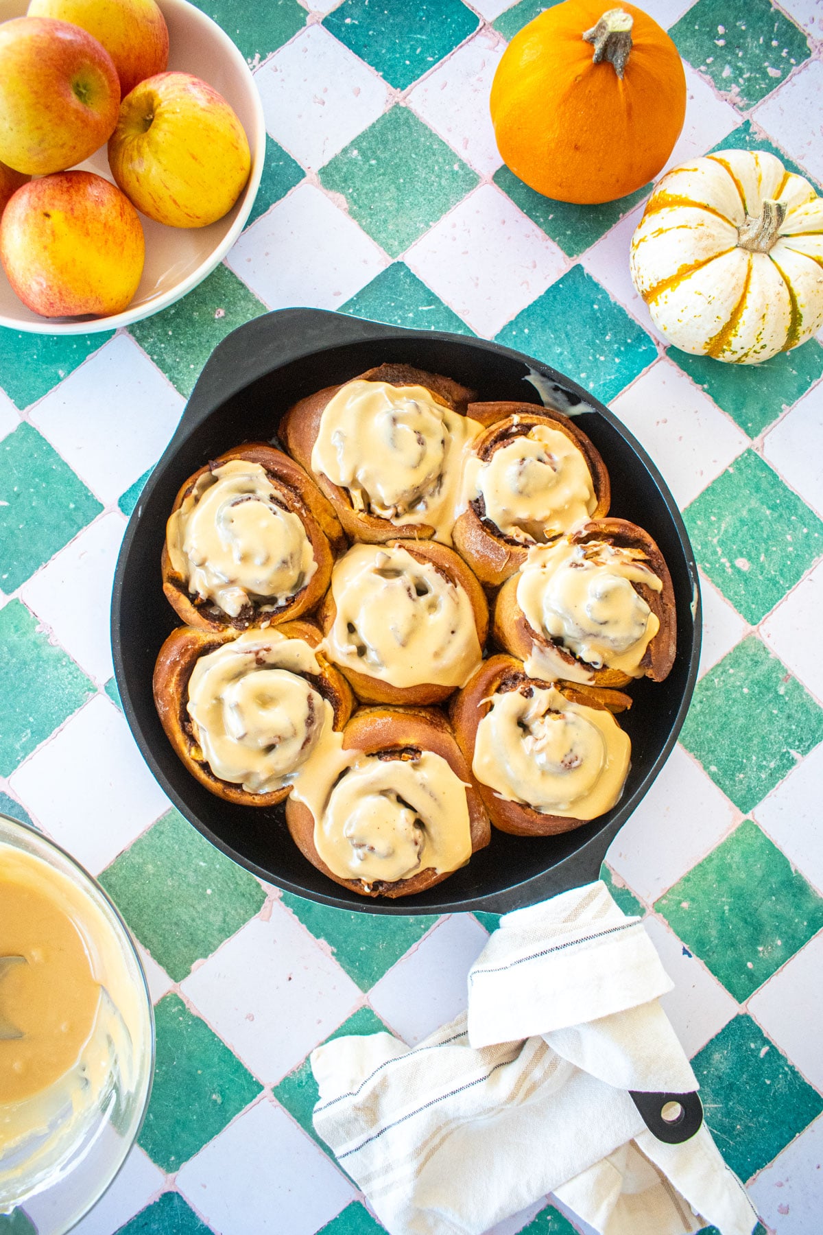 caramel apple pie cinnamon rolls in a cast iron skillet on a tile table with apples.