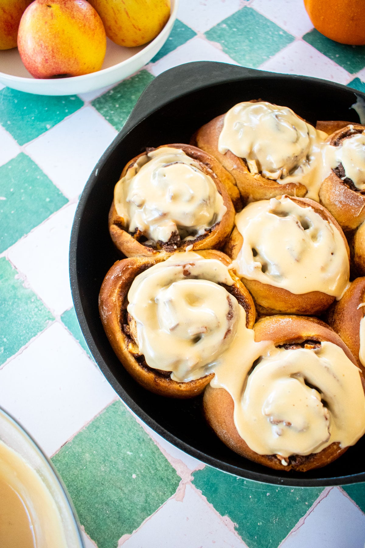 caramel apple pie cinnamon rolls with caramel glaze in a cast iron skillet on a tile table with apples.