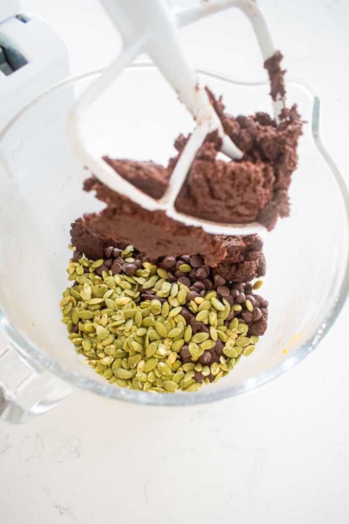 pumpkin chocolate cookie dough with chocolate chips and pumpkin seeds being added to the glass stand mixing bowl.