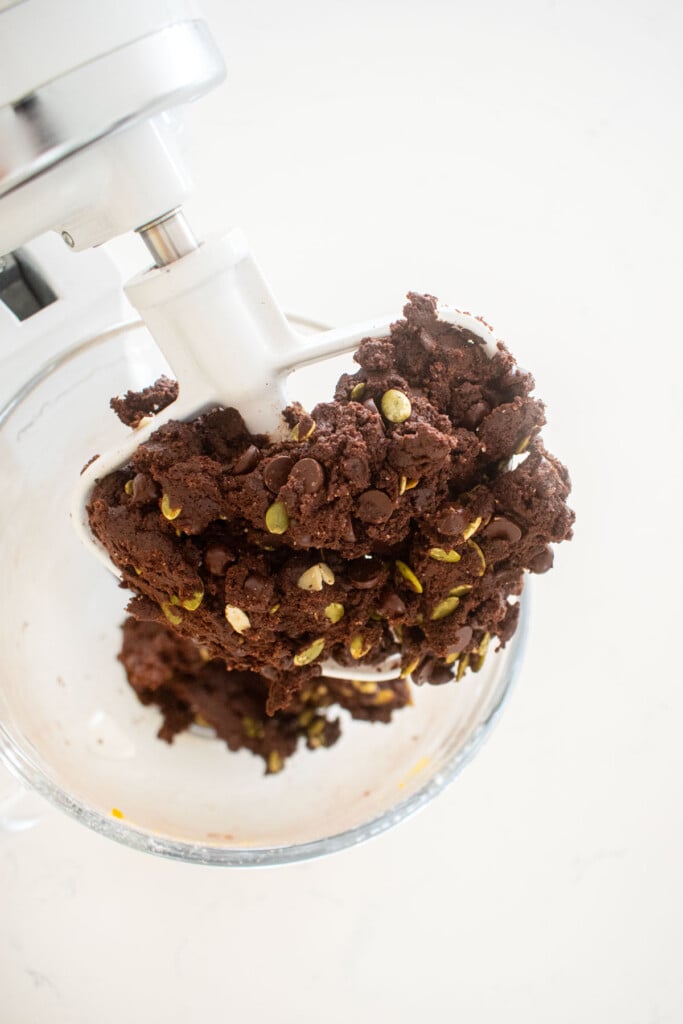 chocolate pumpkin cookie dough with chocolate chips and pumpkin seeds mixed in a glass bowl of a stand mixer.