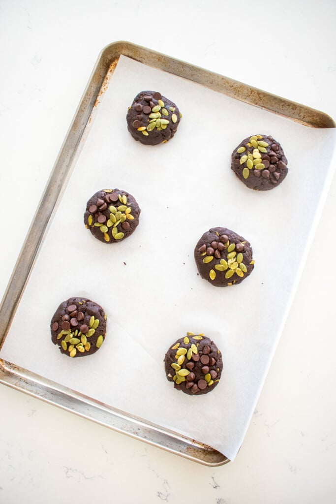 baking sheet with double chocolate pumpkin cookie dough balls and chocolate chips and pumpkin seeds pressed on top on a marble counter.