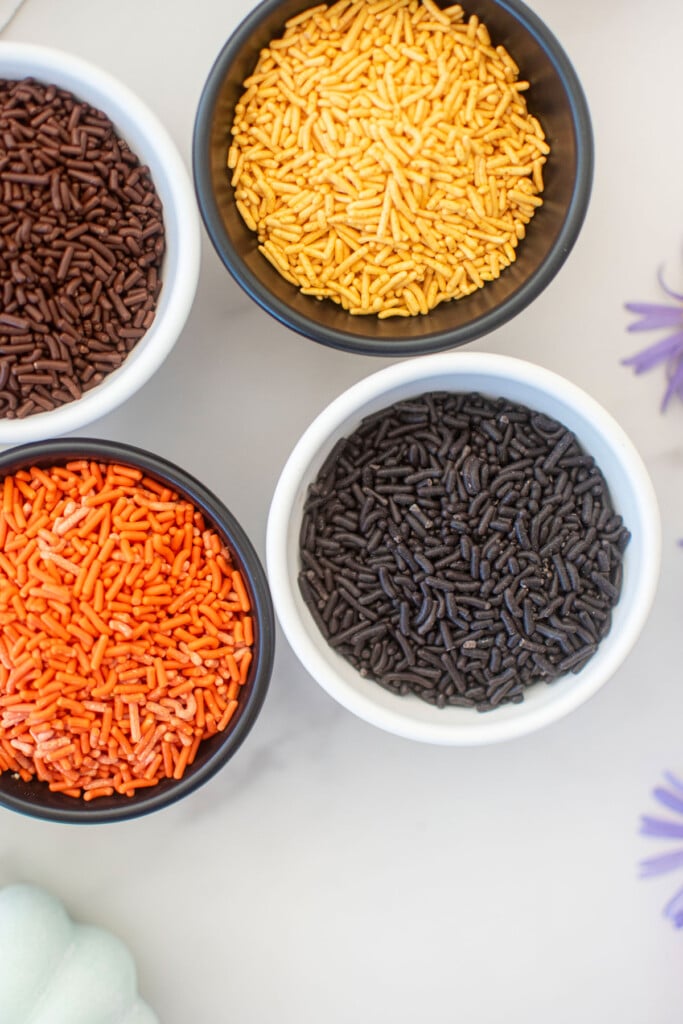 black and orange halloween sprinkles in small bowls on a white marble counter.
