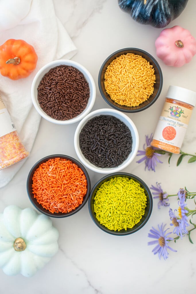 naturally colored halloween sprinkles in small bowls with pumpkins on a white marble counter.