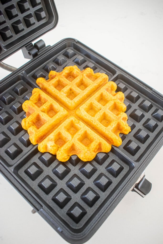 eggless pumpkin waffles in a waffle iron on a white countertop.