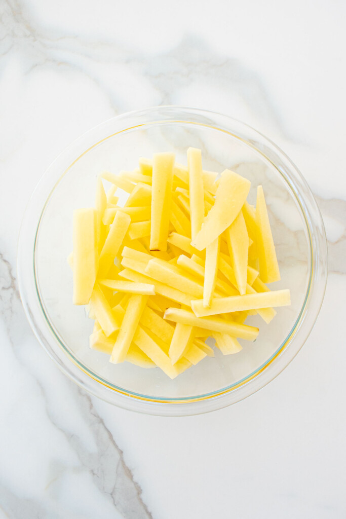 peeled potatoes sliced into fries in a glass mixing bowl on a marble counter.