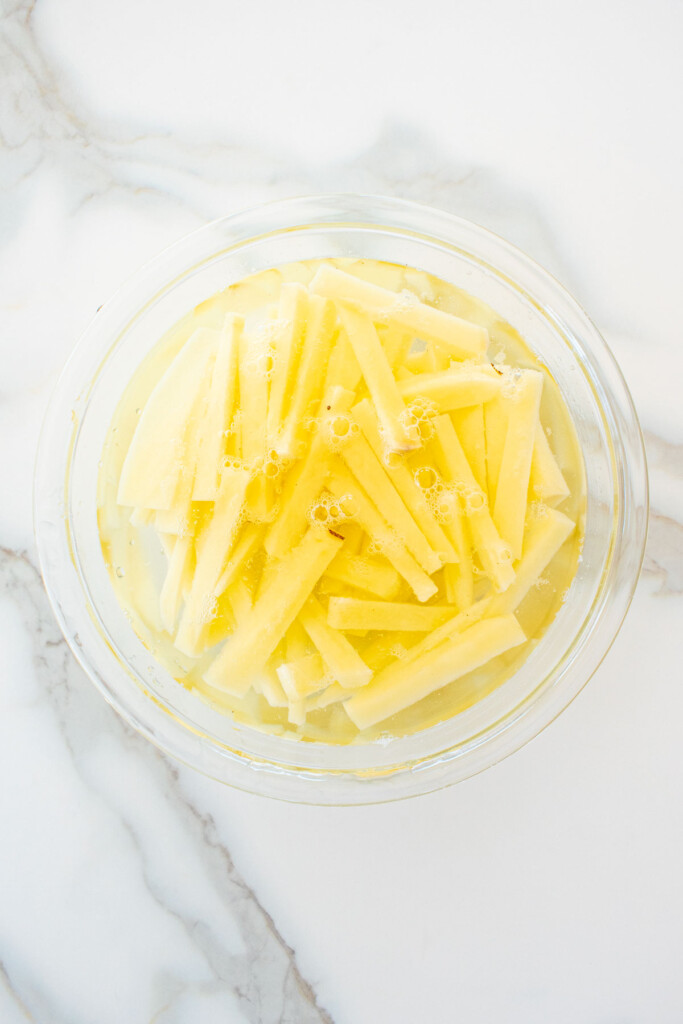peeled potatoes sliced into fries in a glass mixing bowl with water on a marble counter.