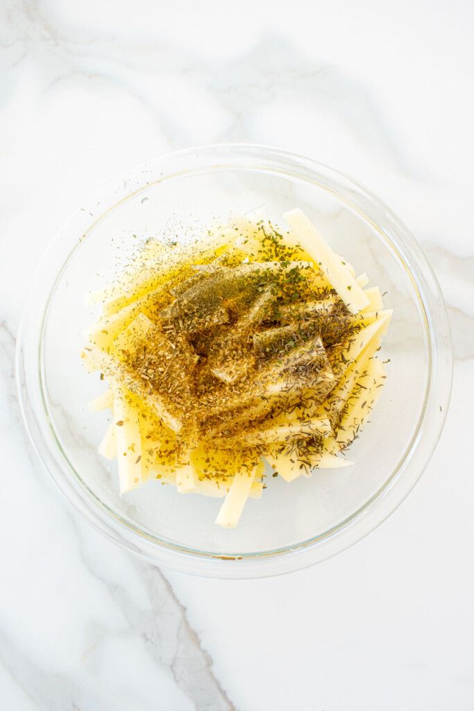 peeled potatoes sliced into fries in a glass mixing bowl on a marble counter with oil and spices.