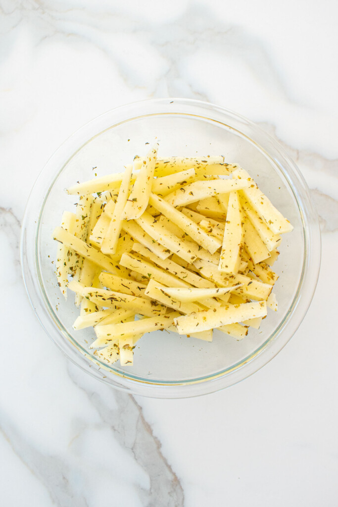 peeled potatoes sliced into fries in a glass mixing bowl on a marble counter with oil and spices.