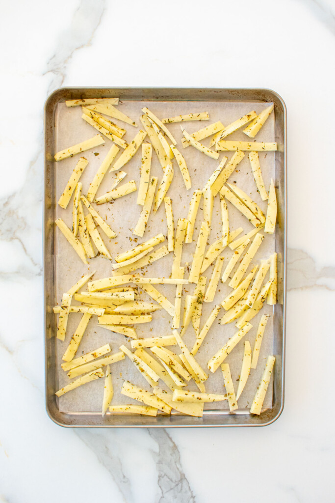peeled potatoes sliced into fries with oil and herbs on a baking sheet on a marble counter.