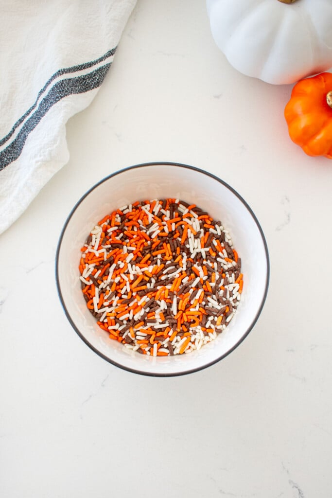 small bowl of mixed orange, black and white sprinkles on a marble counter with mini pumpkins.