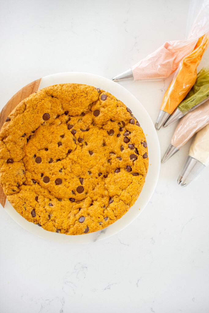 pumpkin chocolate chip cookie cake on a white serving board on a marble counter with frosting bags.