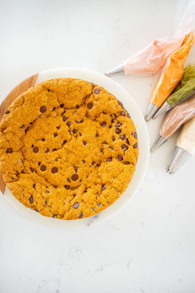 pumpkin chocolate chip cookie cake on a white serving board on a marble counter with frosting bags.