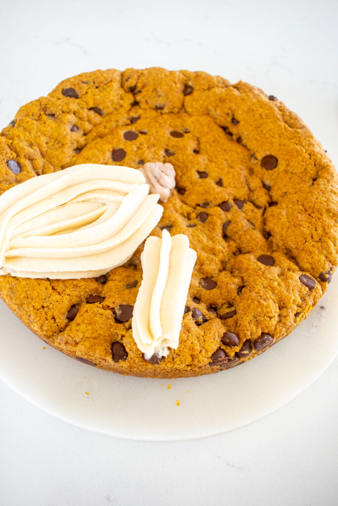 white frosting pumpkins being piped onto a pumpkin chocolate chip cookie cake on a marble counter.