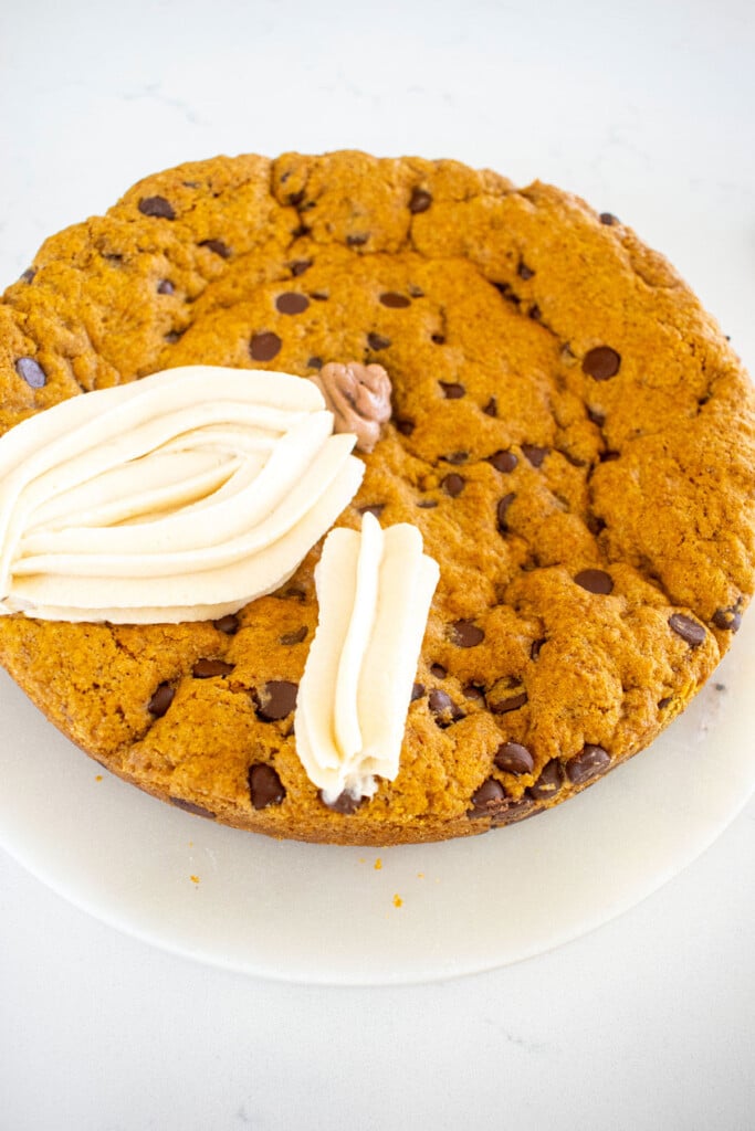 white frosting pumpkins being piped onto a pumpkin chocolate chip cookie cake on a marble counter.