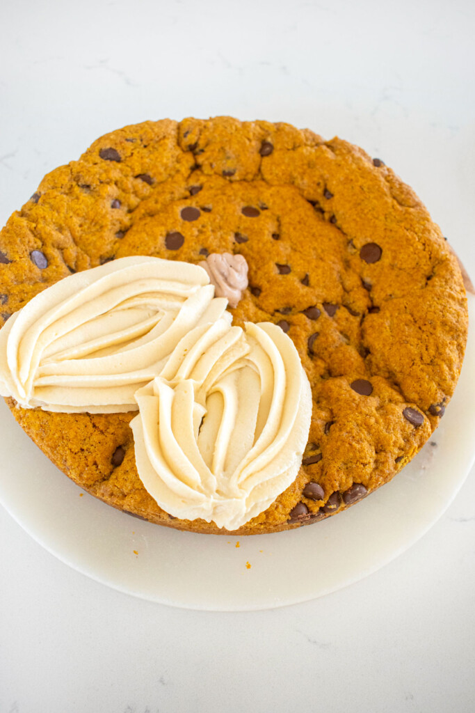 white frosting pumpkins being piped onto a pumpkin chocolate chip cookie cake on a marble counter.