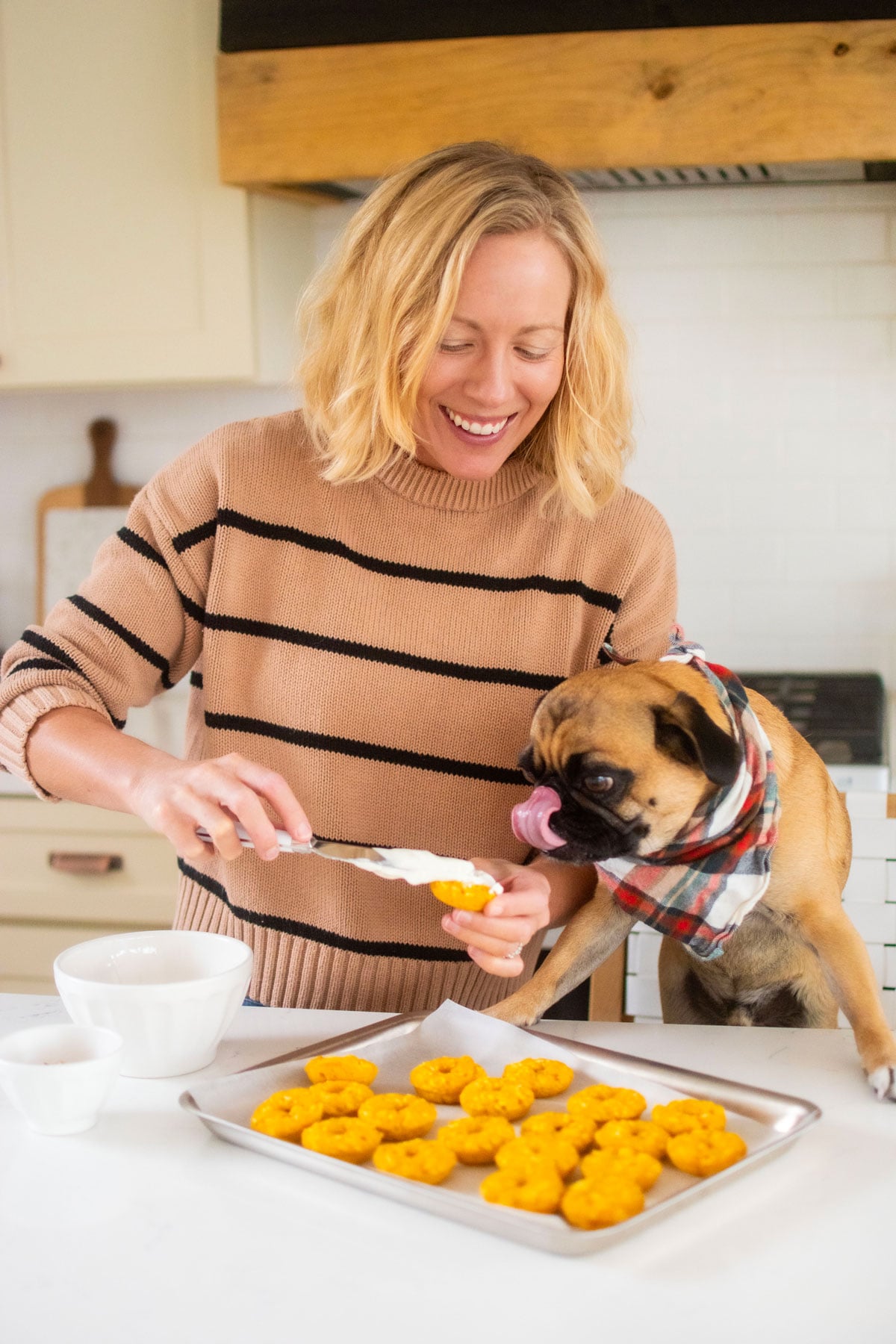 woman and pug in the kitchen frosting pumpkin donuts for dogs.