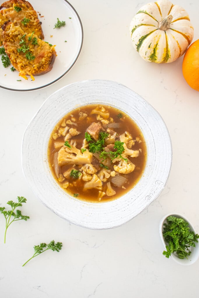 big bowl of slow cooker pumpkin stew with fresh parsley on top on a marble counter with a plate of garlic bread and tiny pumpkins.