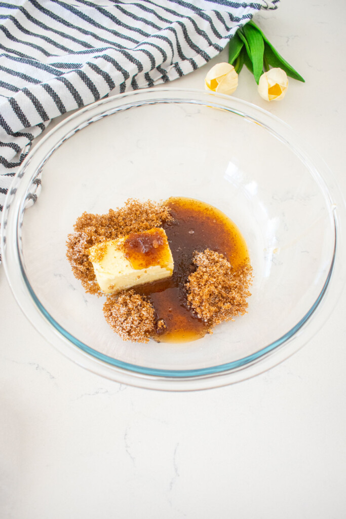 glass mixing bowl with butter, light brown sugar and maple syrup on a marble counter with a kitchen towel and tulips.