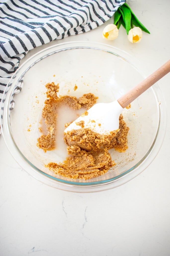 glass mixing bowl with mixed butter, light brown sugar and maple syrup with a spatula on a marble counter with a kitchen towel and tulips.