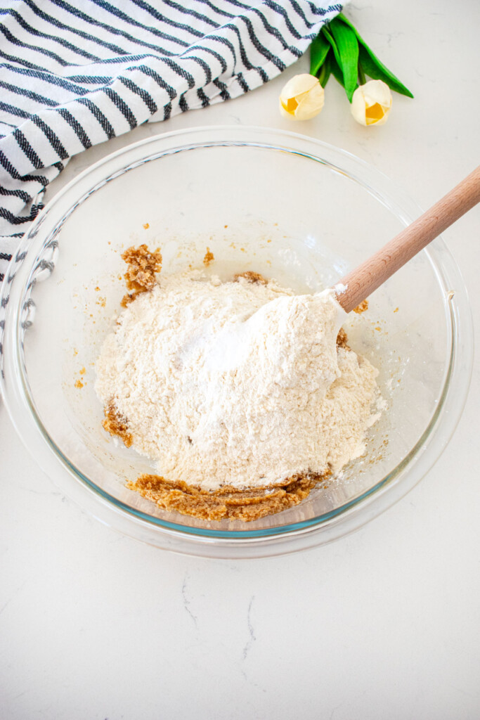 glass mixing bowl with creamed butter and sugar with flour added on top with a spatula in it on a marble counter with a kitchen towel and tulips.
