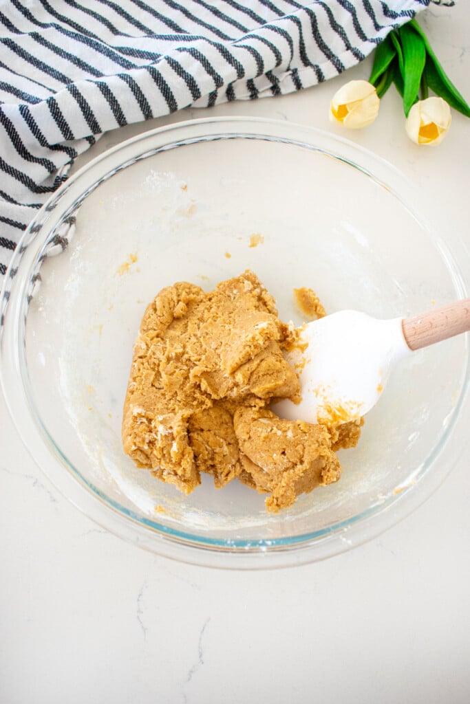 stiff cookie dough in a glass mixing bowl with a spatula on a marble counter with a kitchen towel and tulips.