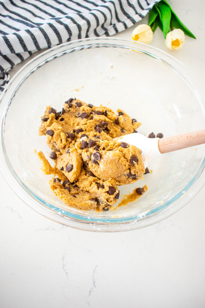 maple chocolate chip cookie dough in a glass mixing bowl with a spatula on a marble counter with a kitchen towel and tulips.