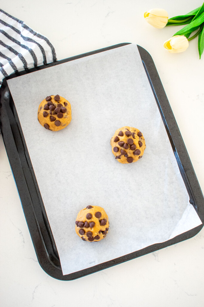 small baking sheet with parchment paper and 3 cookie dough balls on a marble counter with a kitchen towel and tulips.