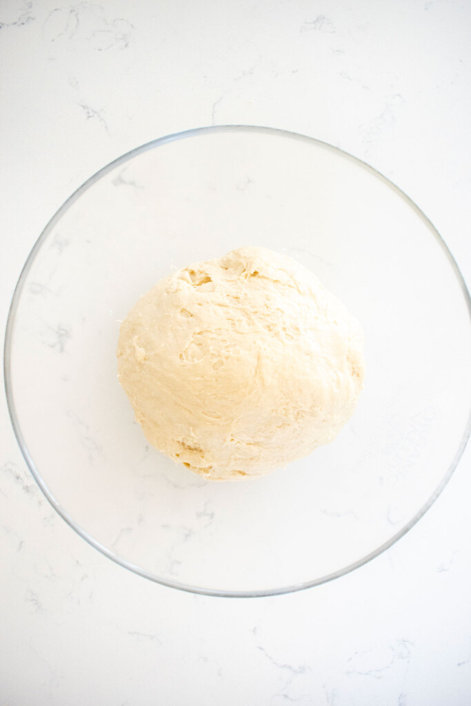 sourdough pretzel bun dough in a glass bowl on a white marble counter proofing.