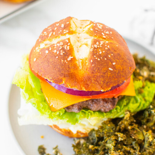 sourdough pretzel bun with a burger and kale chips on a gray plate on a white marble counter.