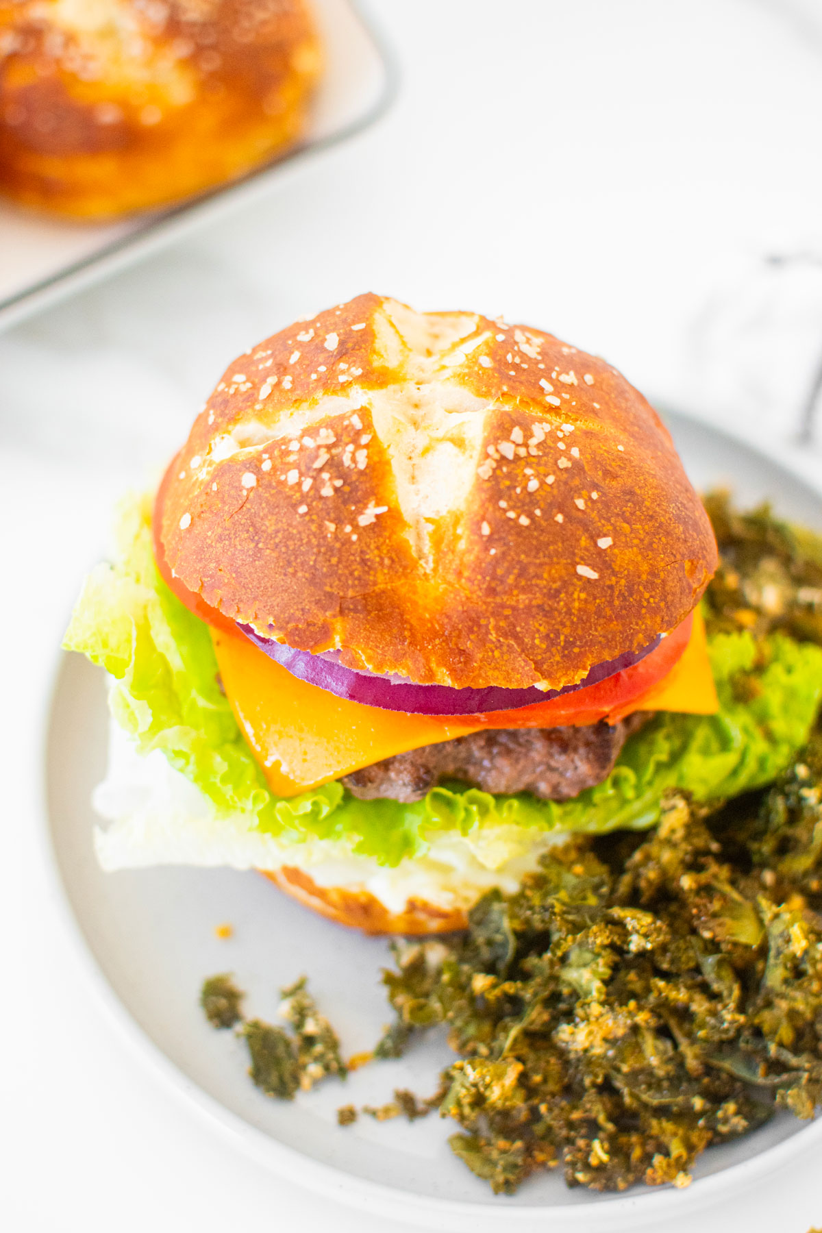 sourdough pretzel bun with a burger and kale chips on a gray plate on a white marble counter.