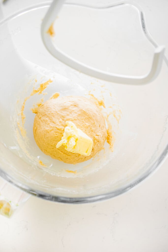 sourdough pretzel bun dough in a glass bowl of a stand mixer with butter being added to it.