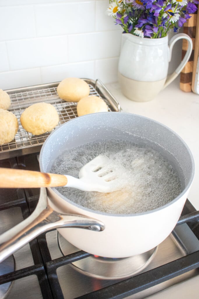 sourdough pretzel buns being submerged in baking soda bath in a pot on the stovetop.