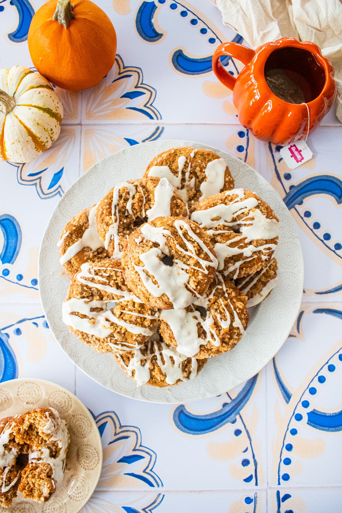 plate of baked chai donuts with glaze on a tile table with pumpkins and a pumpkin mug of tea.