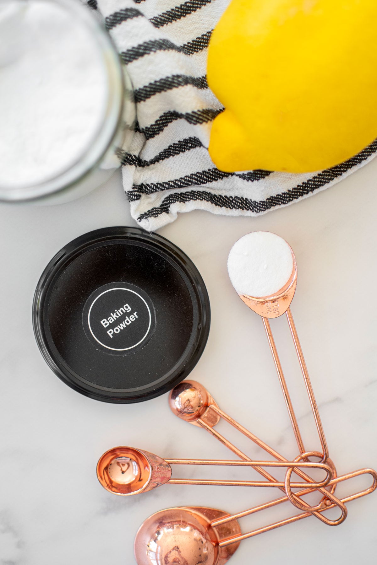black jar of baking powder with label and a teaspoon of baking powder on a white marble counter with a fresh lemon.