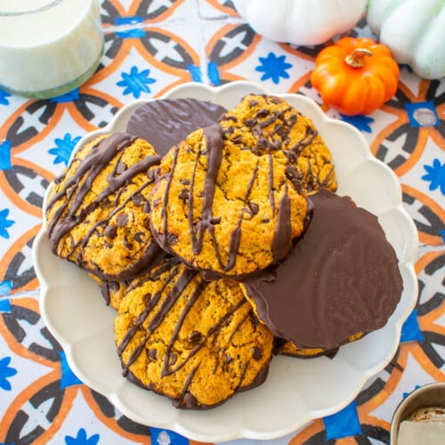 ruffle plate with chocolate dipped pumpkin cookies piled high on a tile table with a glass of milk and pumpkins.