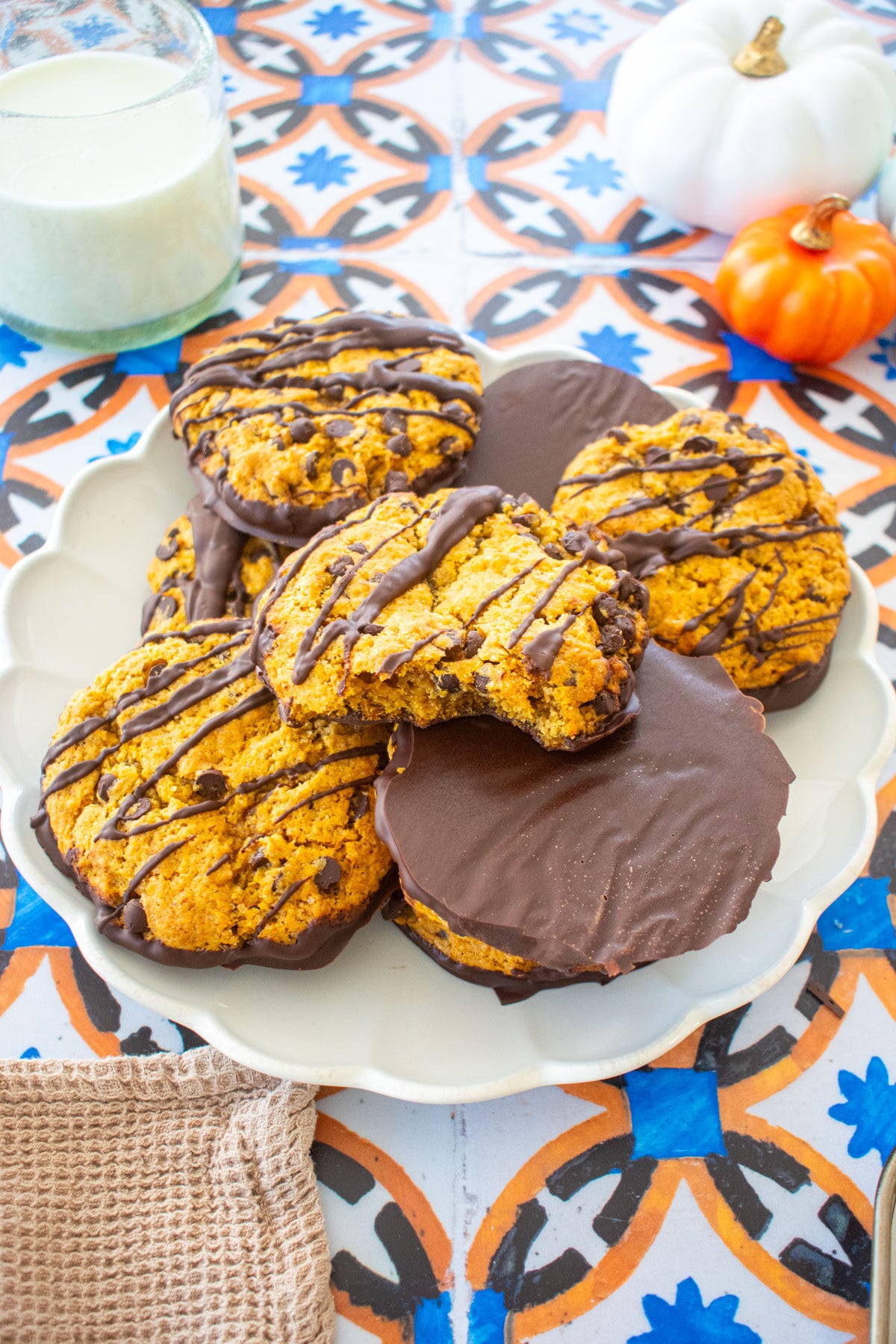 ruffle plate with chocolate dipped pumpkin cookies piled high on a tile table with a glass of milk and pumpkins.