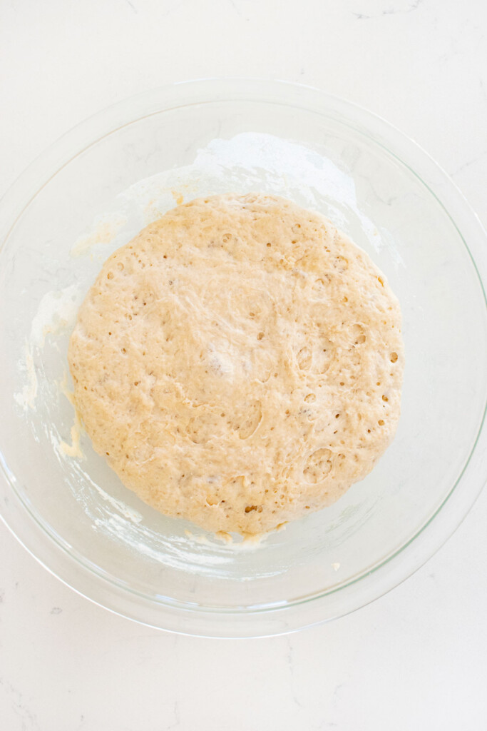 pan pizza dough in a glass mixing bowl on a white marble counter.