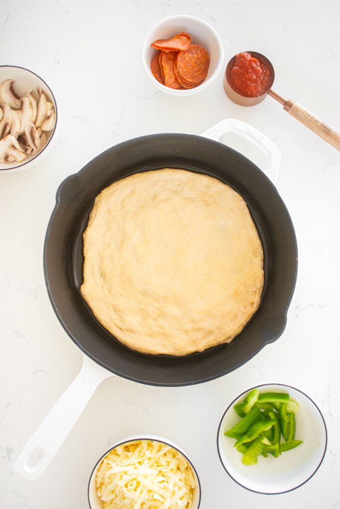 pan pizza dough spread out in a cast iron skillet on a marble counter with pizza toppings around it.