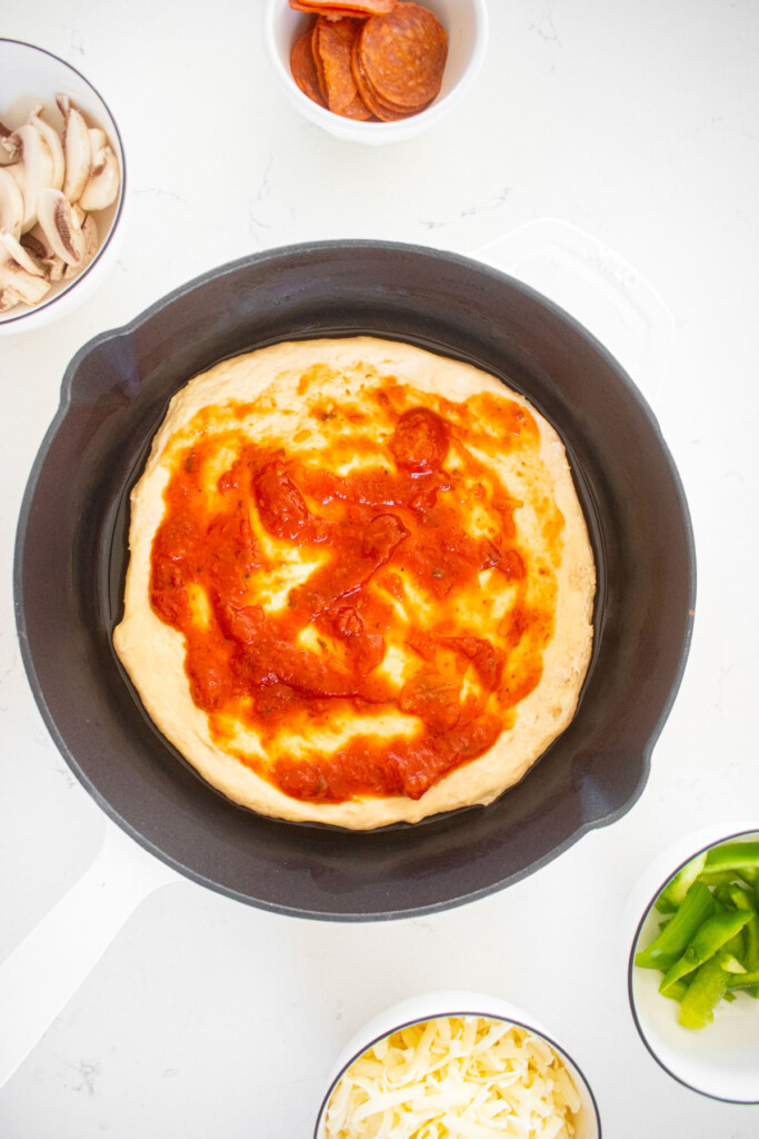 pan pizza being assembled in a cast iron skillet on a white marble counter.