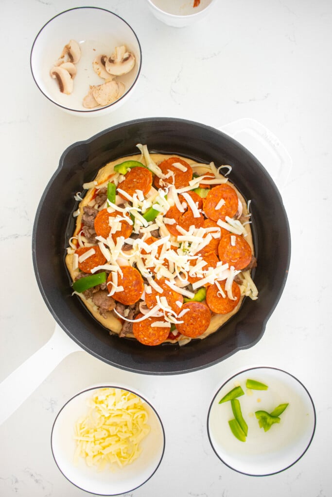 pan pizza being assembled in a cast iron skillet on a white marble counter.