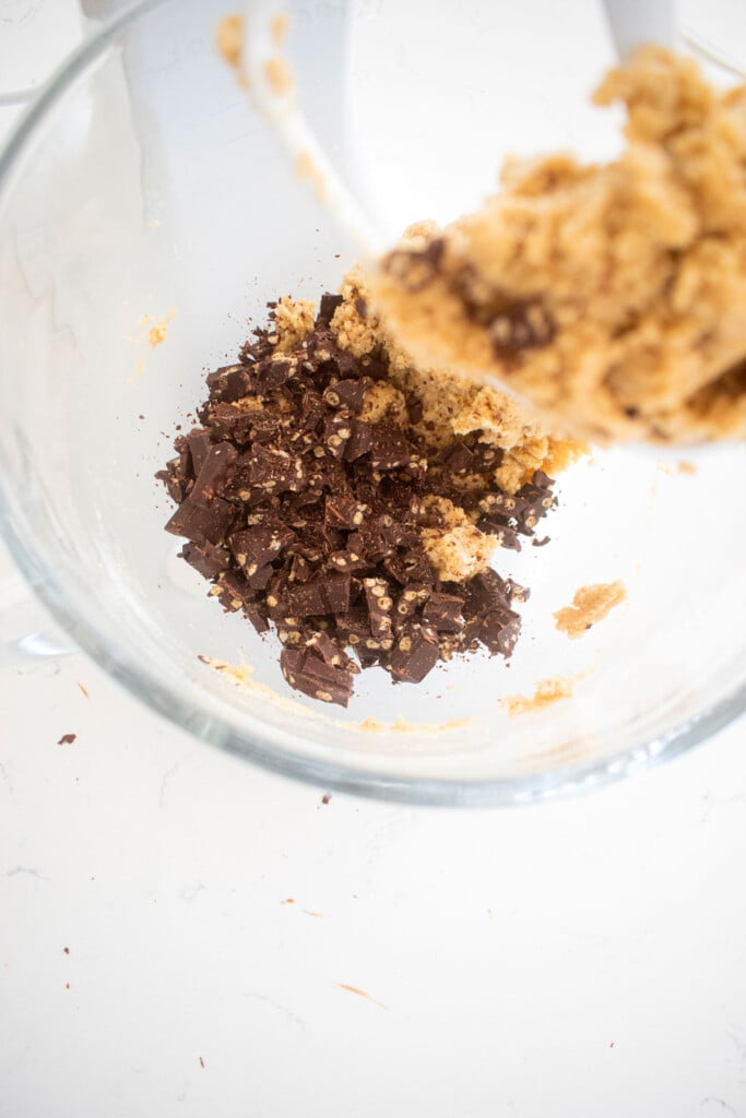 crunch bar cookie dough with chopped up crunch bars being added to the glass mixing bowl on a white marble counter.