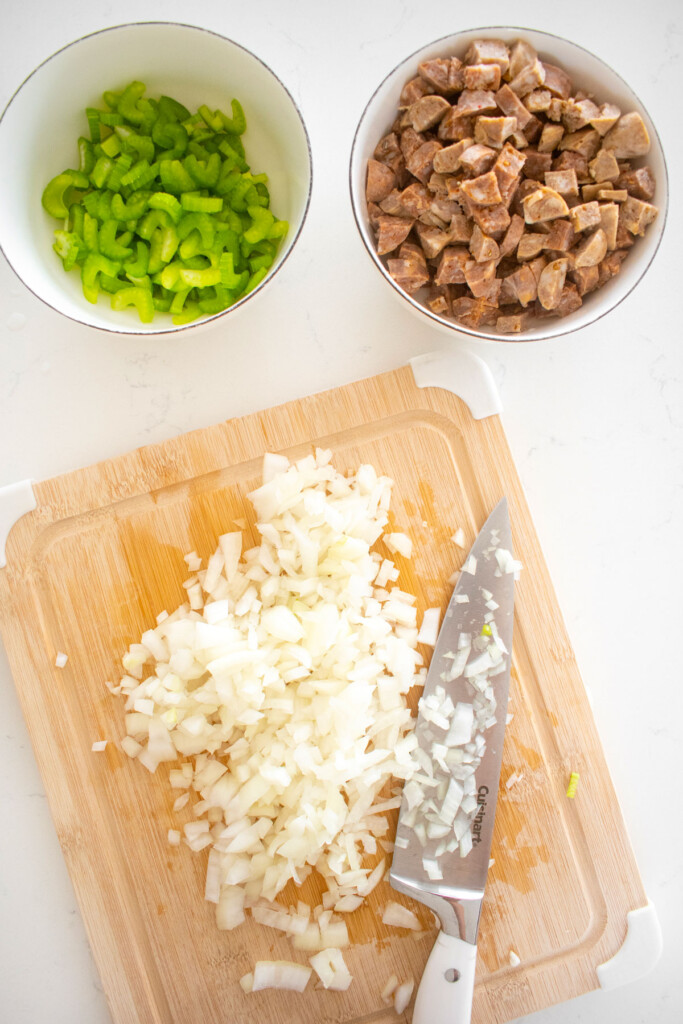 copped onion on a wood cutting board with a bowl of diced italian sausage and celery.