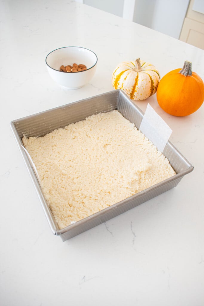 square baking pan with coconut filling pressed into it on a marble counter with tiny pumpkins and a bowl of almonds.