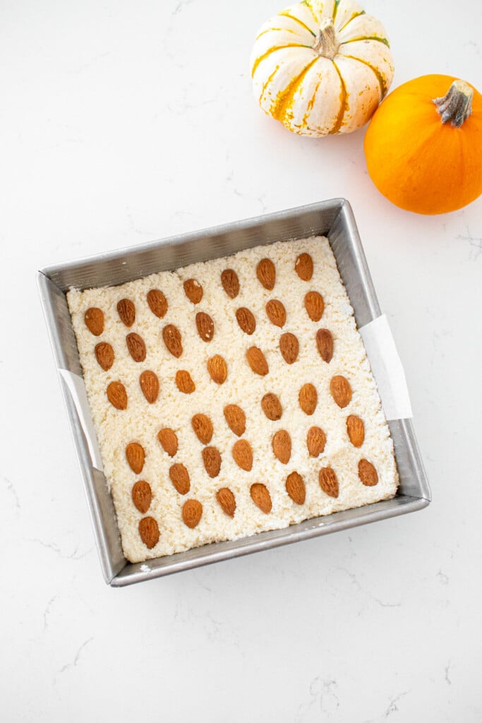 square baking pan with coconut filling and almonds inside on a marble counter with tiny pumpkins.